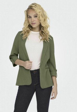 Blazer verde oliva con textura, de corte abierto y mangas tres cuartos. Combinado con una camisa blanca y jeans oscuros.