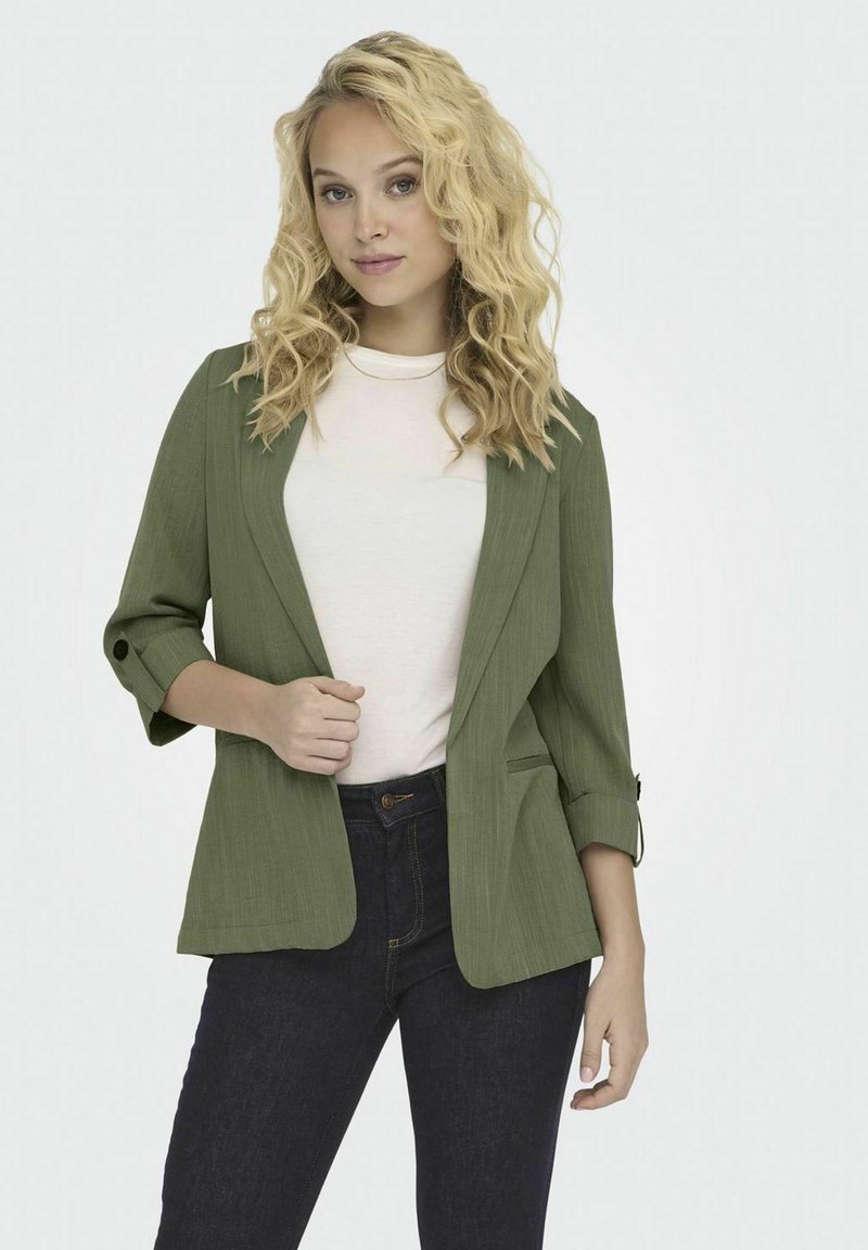 Blazer verde oliva con finitura texturizzata, apertura frontale e maniche a tre quarti. Abbinato a una camicia bianca e jeans scuri.