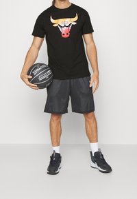 Černé tričko s grafikou býka, šedé síťované šortky a černé sportovní boty. Basketbal míč v levé ruce, stojící na neutrálním pozadí.