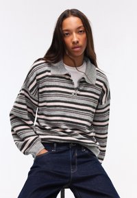 Giovane donna con capelli lunghi, indossa un maglione a righe grigio e rosa e jeans blu scuro, seduta con le mani nelle tasche su uno sfondo bianco.