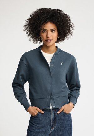 Donna con capelli ricci che indossa un cardigan blu scuro con bottoni sopra una camicia bianca e jeans blu, mani nelle tasche, sfondo neutro.