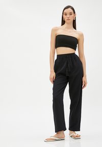 Svart ribbad crop-top, lösa svarta byxor med dragsko och fransar, och vita flip-flops. Slät textur, avslappnad passform, minimalistisk design.
