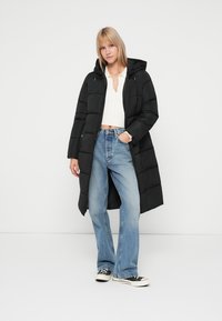 Črna puffer jakna s kapuco, dolge in strukturirane oblike ter kviltano teksturo, v kombinaciji z svetlo modrimi širokimi kavbojkami in črnimi supergami.