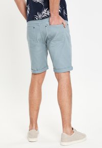 Pantalones cortos celestes con puños enrollados, bolsillos traseros y una textura suave, combinados con zapatillas deportivas gris claro y una camisa de patrón oscuro.
