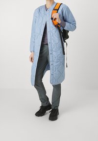 Ljusblå quiltad lång kappa med rund halsringning, tryckknappar och lös passform. Kombinerad med mörka byxor och en svart ryggsäck med orange axelrem.