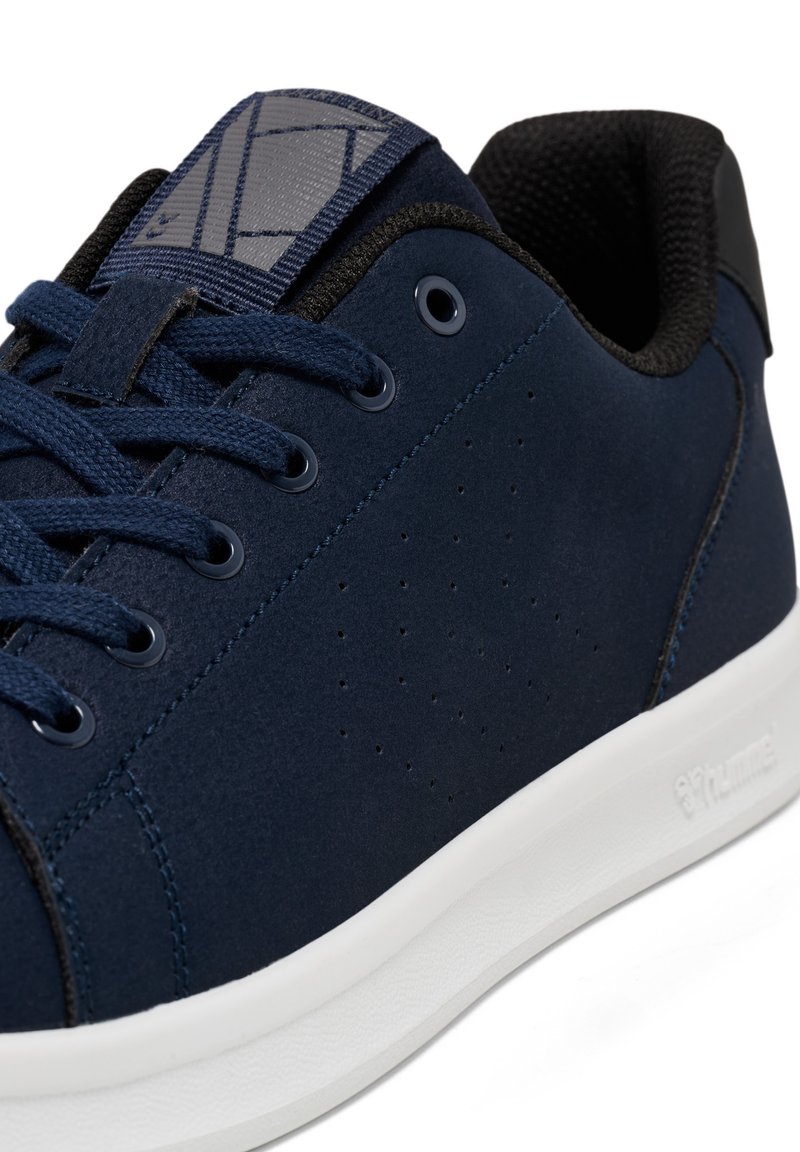 Zapatilla azul marino con parte superior de ante, detalles perforados y cuello de tela negra texturizada. Presenta cordones planos en azul marino y una suela de goma blanca.