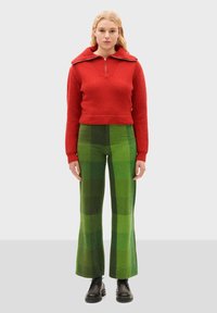 Pull en tricot rouge avec un col zippé et large, associé à un pantalon évasé à carreaux verts, et des bottes noires à lacets épaisses.