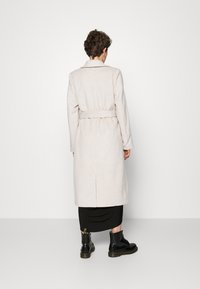Abrigo de lana beige con cintura ceñida, mangas largas y abertura trasera, combinado con una falda negra y botas negras con cordones.