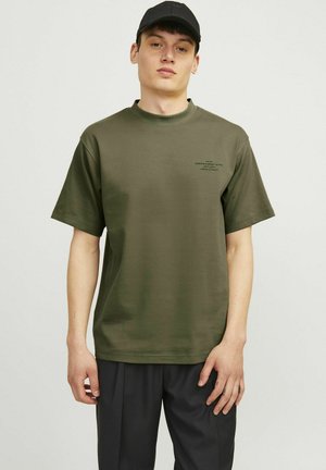Camiseta de algodón verde oliva de gran tamaño con mangas cortas, escote redondo y un pequeño detalle impreso en el pecho, combinada con pantalones oscuros.