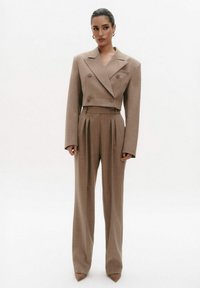 Bruin op maat gemaakt pak met een cropped dubbele jas en hoge taille, geplooide broek; gladde stof, brede mouwen en puntige hakken.