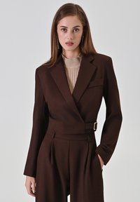 Bruine op maat gemaakte blazer met een aangesloten ontwerp, een borstzakje en een gespdetail. Gecombineerd met een lichtbeige geribd topje.