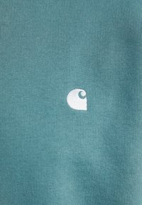 Tyrkysová látka s jemne ryhovanou textúrou, ktorá má biele vyšívané logo v tvare štýlizovaného písmena "C" v dolnom pravom rohu.