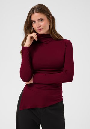 Femme aux longs cheveux bruns portant des boucles d'oreilles créoles dorées et un haut col roulé rouge foncé ajusté, posant avec une main près du menton.