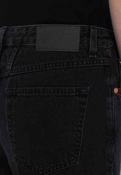 Sorte denimjeans med teksturert finish, som har en rektangulær lærpatch merket "CLOSED" på baksiden av livet og subtile kobberdetaljer.