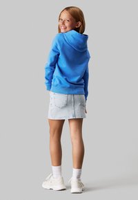 Sudadera azul con capucha y corte relajado, combinada con una falda de mezclilla clara. Zapatillas blancas y calcetines hasta el tobillo completan el conjunto.