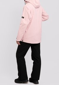 Giacca impermeabile rosa con cappuccio, dal taglio comodo e tasche laterali con zip, abbinata a pantaloni neri e calzature isolate.