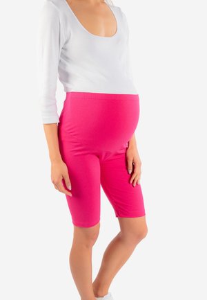 Kobieta w ciąży ubrana w biały top i jasnoróżowe, ciążowe szorty stojąca na jednolitym tle.