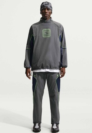 Homme debout face à l'objectif, portant un survêtement gris assorti avec des touches vert fluo et un foulard à motifs, sur un fond uni.