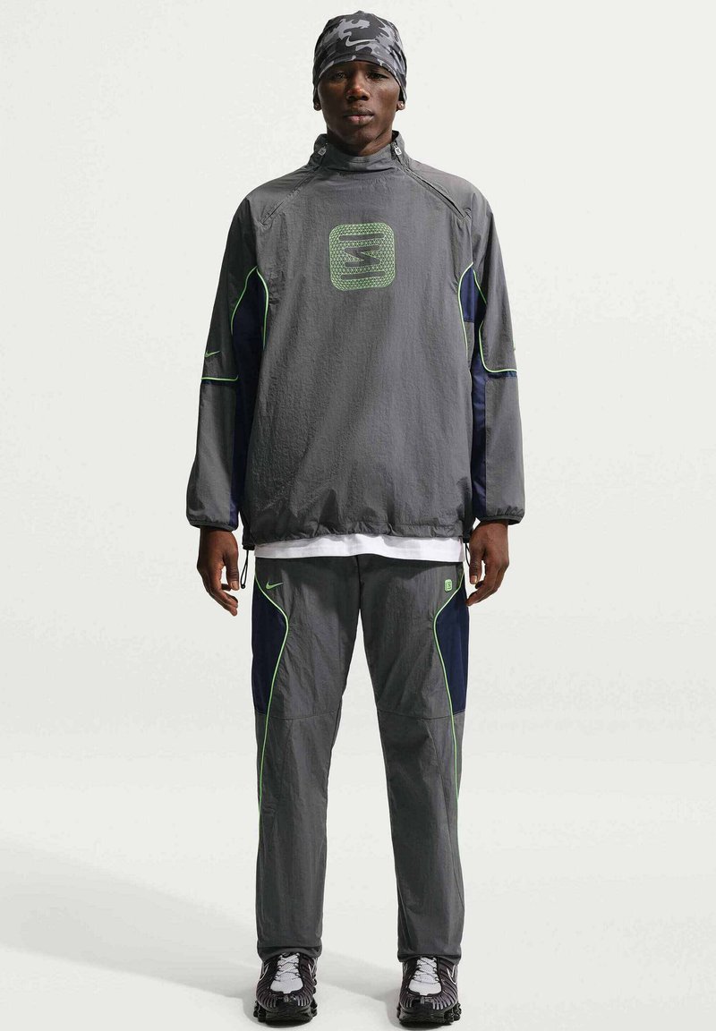 Man staat frontaal, draagt een bijpassend grijs trainingspak met neon groene accenten en een hoofddoek met patroon, tegen een effen achtergrond.