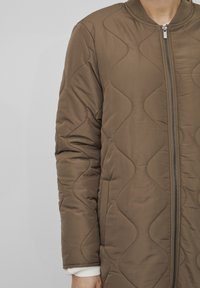 Brun vadderad jacka med dragkedja, ribbad krage och en slät yta med en vågig sömnadsstil. Vita manschetter synliga.