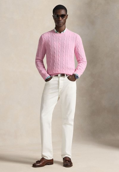 Homme portant des lunettes de soleil, pull rose en maille torsadée par-dessus une chemise à motifs, pantalon blanc, mocassins en cuir marron, mains dans les poches, debout devant un fond neutre.