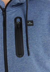 Felpa blu con tessuto testurizzato, dotata di una tasca con zip nera e un'etichetta con logo nera. I cordini sono regolabili con punte in plastica.