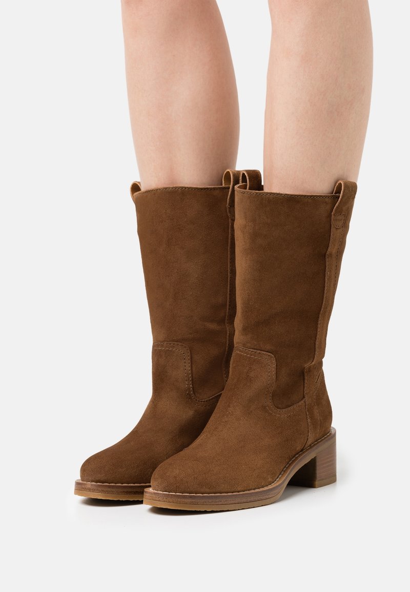 Bottes en suède marron mi-mollet avec bouts arrondis et talons blocs bas portées sur jambes nues sur fond blanc.