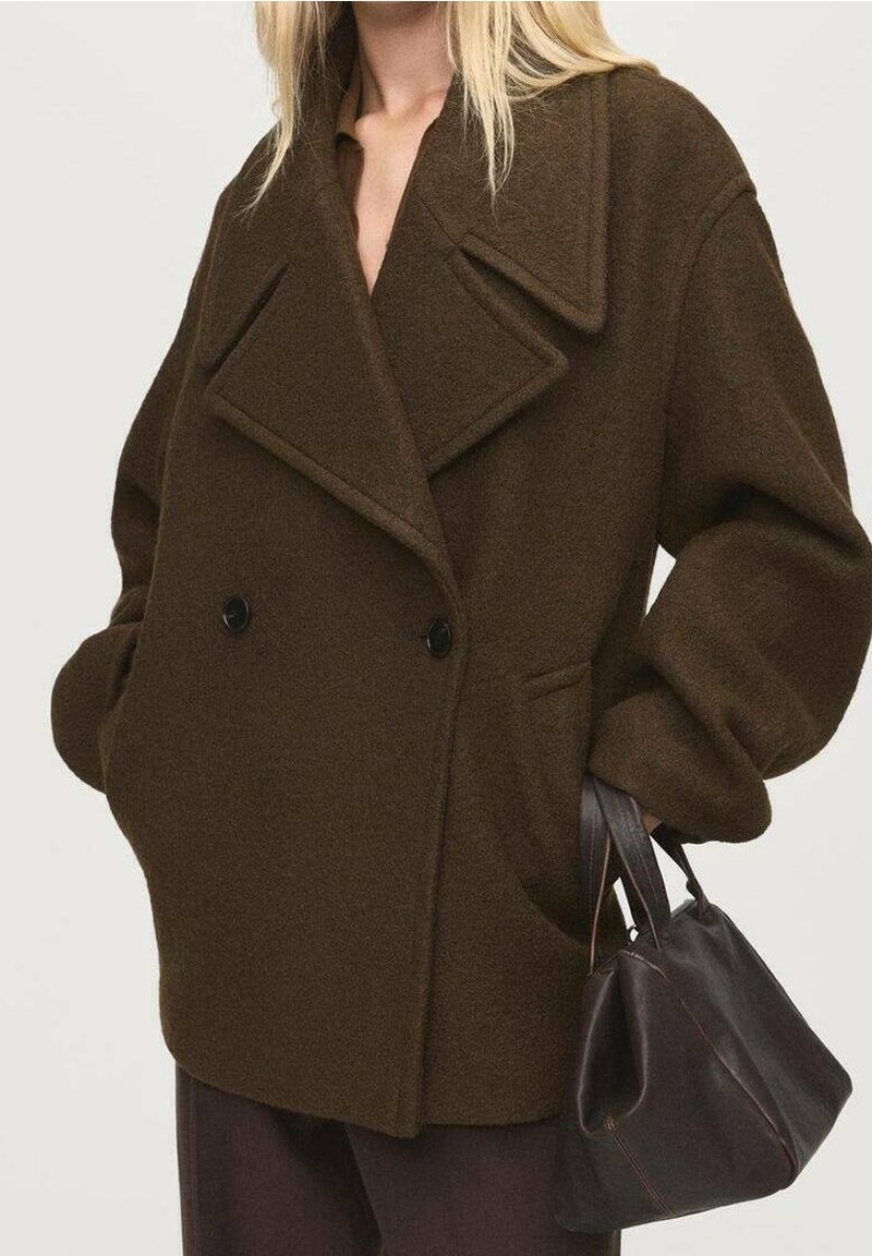 Femme portant un manteau en laine brun foncé oversize avec de grands revers, tenant un sac à main en cuir brun foncé dans une main.