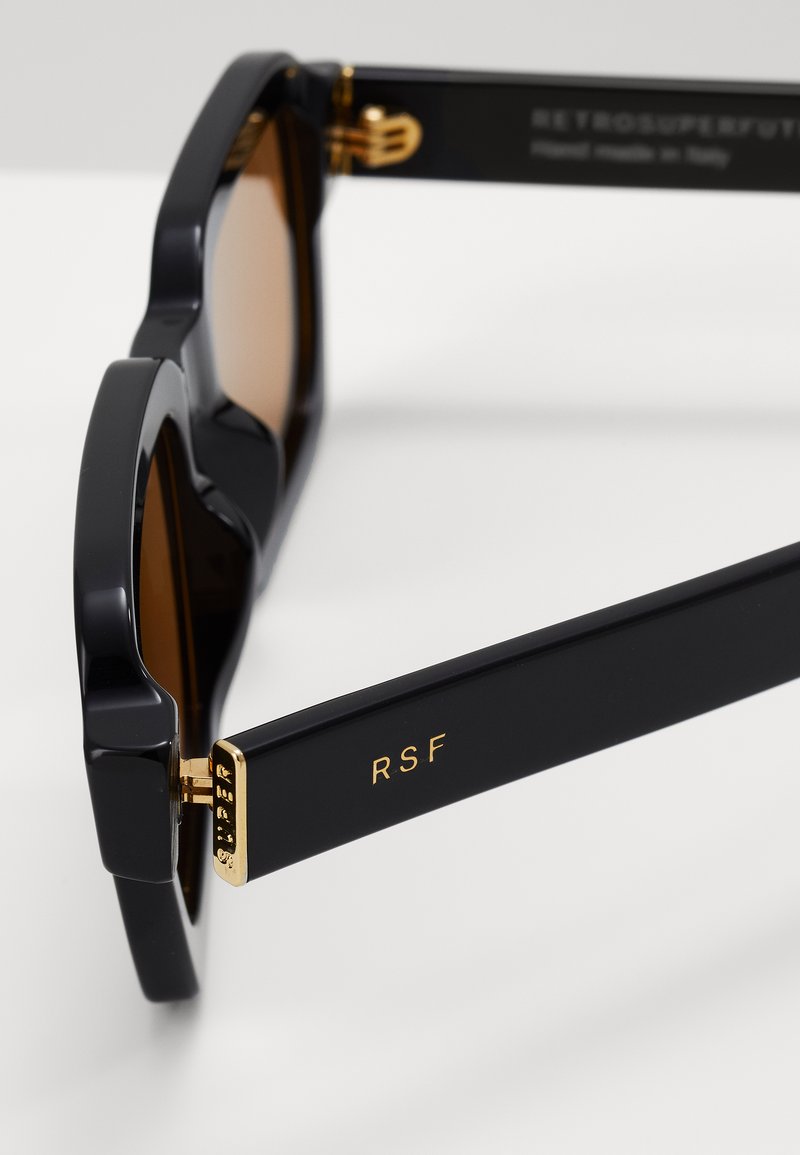 Occhiali da sole neri oversize con lenti marroni. Presentano accenti dorati sulle tempie e un marchio sul braccio. Finitura liscia e lucida.