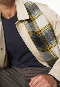 Man som bär en beige jacka, blå tröja, grå byxor och en halsduk i gul, svart och vit rutigt över axeln.