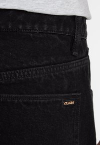 Black denim jeans with a smooth texture; features a small, gold Volcom logo on the back pocket and subtle stitching details.