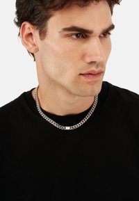 Sølv kædedyse med kraftige led og et tekstureret midtstykke, båret af en person i en sort T-shirt mod en ensfarvet baggrund.