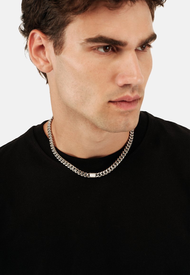 Sølv kædedyse med kraftige led og et tekstureret midtstykke, båret af en person i en sort T-shirt mod en ensfarvet baggrund.