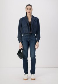 Veste en denim bleu marine coupée avec des boutons croisés, haut noir, jeans bleus et baskets blanches avec des semelles en caoutchouc. Tient un sac noir.