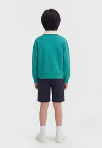 Παιδί που στέκεται με την πλάτη γυρισμένη, φορώντας γαλάζιο μακρυμάνικο μπλουζάκι, σκούρα μπλε σορτς, λευκές κάλτσες και λευκά αθλητικά παπούτσια, πάνω σε απλό φόντο.