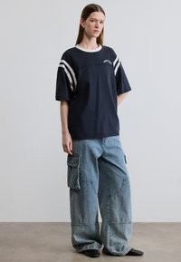 Μπλε ναυτικό oversized t-shirt με λευκές ρίγες στα μανίκια, με στρογγυλή λαιμόκοψη. Συνδυάζεται με παντελόνι cargo μπλε με φαρδιές ίσιες γραμμές. Υφή υφάσματος με ανάγλυφη επιφάνεια.