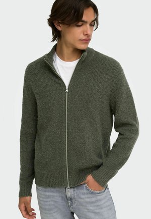 Homme portant un pull zippé texturé vert foncé sur un t-shirt blanc, avec un jean bleu clair, regardant vers le bas sur sa gauche.