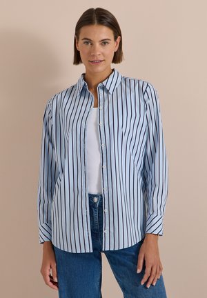 Femme portant une chemise bleu clair à rayures verticales boutonnée sur un haut blanc et un jean bleu, debout devant un fond beige uni.