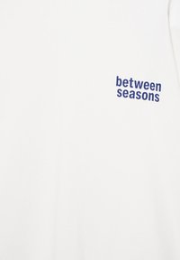 Tissu blanc avec le texte bleu marine « between seasons » imprimé en haut à droite.