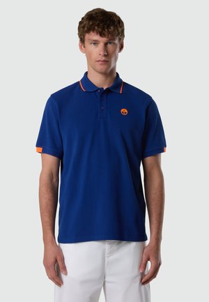 Uomo che indossa una polo a maniche corte blu con rifiniture e emblema arancioni, abbinata a pantaloni bianchi, in piedi davanti a uno sfondo semplice.