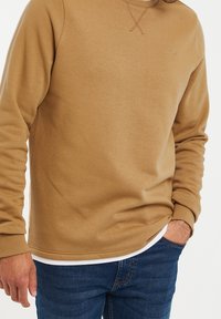 Sudadera marrón claro con cuello redondo, puños y dobladillo acanalados, y un pequeño logo en el pecho, llevada sobre una camisa blanca, combinada con jeans azules.