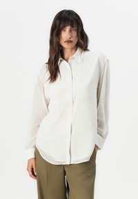 Long-sleeve, button-up shirt in soft, light cream fabric. Features a chest pocket and relaxed fit, with a smooth, slightly glossy texture.