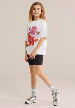 Jeune fille portant un T-shirt blanc oversize avec des fleurs colorées et le texte « Flower Days », un short noir, des chaussettes grises et des baskets blanches, souriante.
