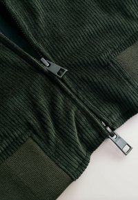 Donkergroene zip-up jas van ribbelstof. Beschikt over een zwarte rits en een effen groene panel bij de mouwen en de zoom.