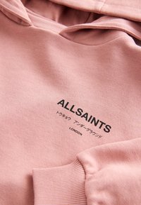 Ružová mikina s čiernym logom ALLSAINTS, japonskými znakmi a textom "LONDON" na hrudi, ukazujúca rebrované manžety a detaily kapucne.