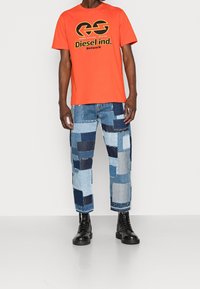 Oranges Baumwoll-T-Shirt mit schwarzem Grafiklogo; Patchwork-Jeans in hellblauen und dunkelblauen Farbtönen; schwarze Schnürstiefel.