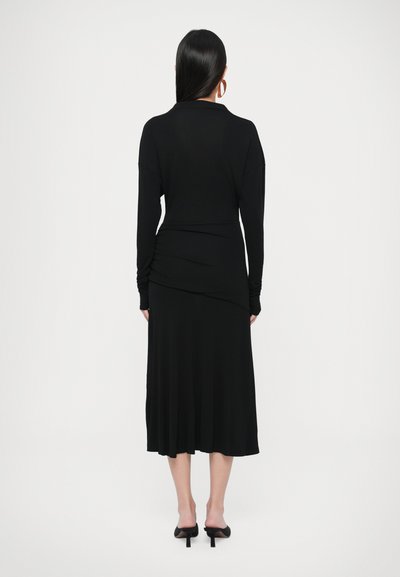 Fekete, hosszú ujjú ruha, testhez álló felsőrésszel, redőzött részletekkel és bővülő szoknyával. Sima anyag, részben a deréknál összehúzva.