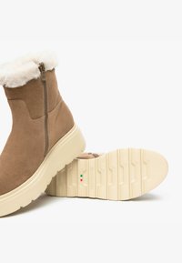 Bottines en daim beige avec un col en fausse fourrure blanche, dotées d'une fermeture éclair sur le côté et d'une semelle en caoutchouc texturé de couleur crème.
