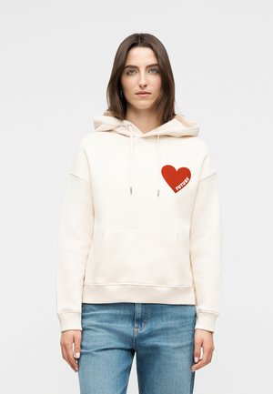 Femme aux cheveux bruns mi-longs portant un sweat à capuche crème avec un cœur rouge et le mot « FUTURE » sur la poitrine, ainsi qu'un jean bleu.