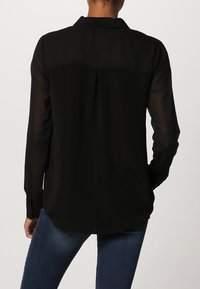 Black, long-sleeve shirt with a collar, made of lightweight, sheer fabric. Features a back yoke and rounded hem, paired with denim jeans.